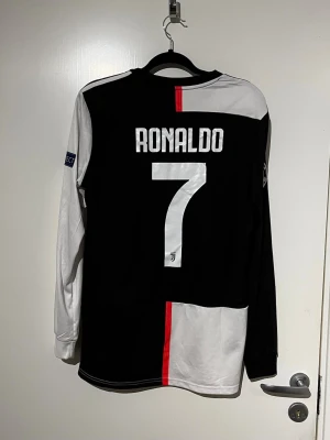 Juventus Ronaldo matchtröja långärmad - Juventus långärmad matchtröja med Ronaldo 7 på ryggen. Tröjan är svart och vit med röd rand i mitten, Juventus klubbmärke och Jeep-logga på bröstet samt Champions League-märke på ärmen. Tillverkad i funktionsmaterial, perfekt för fotboll.