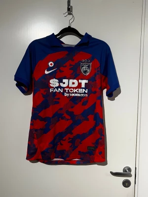 Blå och röd fotbollströja Nike - Säljer en snygg fotbollströja från Nike i blått och rött med kamouflagemönster. Tröjan har korta ärmar, klubbmärke på bröstet och tryck med 'JDT FAN TOKEN'. Perfekt för dig som gillar sportig stil och vill sticka ut på planen.