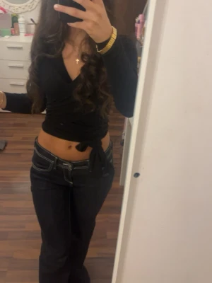 Svart omlottopp med knyt - Säljer en svart omlottopp med långa ärmar och knyt i midjan. Toppen har en v-ringad urringning och croppad passform som ger en trendig look. Perfekt att styla med jeans eller kjol för en snygg outfit.
