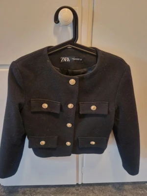 Svart kort jacka från Zara - Stilren svart kort jacka från Zara med rund halsringning och dekorativa guldknappar framtill. Fyra fickor med lock och lång ärm. Jackan har en elegant och klassisk look, perfekt för att lyfta vilken outfit som helst.