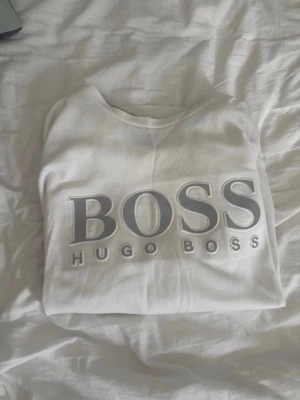 Vit sweatshirt från Hugo Boss M - Snygg vit sweatshirt från Hugo Boss med stort grått BOSS-tryck och HUGO BOSS-text på bröstet. Tröjan har rund halsringning och långa ärmar. Perfekt för en clean och stilren look. Har också en i S
