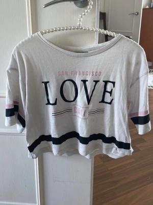 Vit crop top med tryck LOVE - Vit crop top med texten 'SAN FRANCISCO LOVE CALI' i svart och rosa. T-shirten har breda svarta och rosa ränder på ärmarna och nedtill. Rund halsringning och kortare passform. Perfekt för en avslappnad och trendig look.