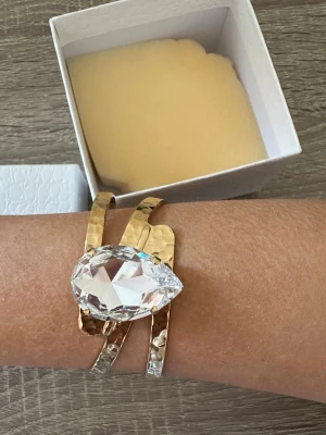 Guldfärgad armband med stor kristall från Caroline svedbom - Säljer en elegant guldfärgat armband från Caroline Svedbom med en stor, klar kristall i droppform. Armbandet har en bred, hamrad design och levereras i original ask. Perfekt för dig som vill sticka ut med ett exklusivt smycke. Armbandet är 18cm i diameter och aldrig använd! OBS: FRAKTAR INOM 24H!🩷