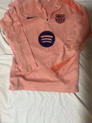Rosa FC Barcelona träningströja Nike - Snygg långärmad träningströja från Nike i ljusrosa med FC Barcelona-logga och Spotify-tryck på bröstet. Tröjan har halv dragkedja framtill och orange detaljer på ärmarna. Perfekt för fotbollsträning eller som sportig vardagsstil.