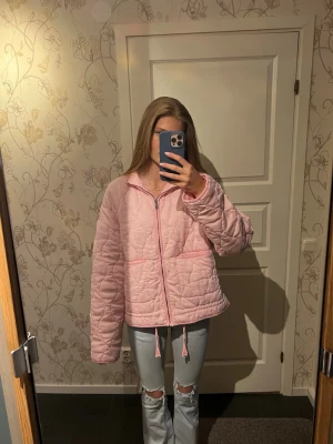 Ljusrosa quiltad jacka med dragkedja - Quiltad jacka i ljusrosa med dragkedja framtill och dragsko i nederkant. Jackan har två stora fickor och en avslappnad passform. Perfekt för lager-på-lager och ger en trendig vibe till din outfit. Helt ny och aldrig använd bara provad.