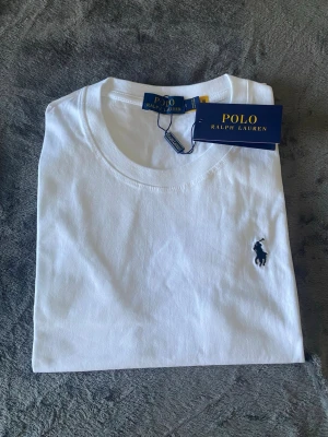 Ralph Lauren T-shirt  - Klassisk vit t-shirt från Polo Ralph Lauren med rund halsringning och den ikoniska mörkblå logotypen broderad på bröstet. Tillverkad i mjuk bomull för en skön och stilren look. Perfekt basplagg för alla tillfällen.