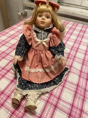 Porslinsdocka med rosa klänning - Charmig porslinsdocka med långt blont hår, blå ögon och en stor rosa rosett. Dockan har en rosa klänning med spetsdetaljer, vit rosett och blommigt mönstrad underklänning. Skorna är vita med snörning och dockan har vita strumpor. Perfekt som samlarobjekt eller dekoration.