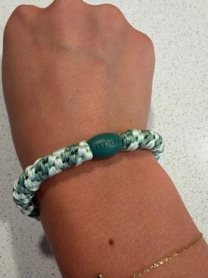 Kknekki hårband  - Snyggt armband i flätad design med gröna och vita toner. Tillverkat av slitstarkt snöre och har en oval grön detalj med texten 'FJÄLLRÄVEN'. Perfekt accessoar för en avslappnad och trendig stil.