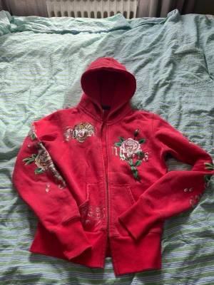 Ed Hardy 2000 - Dragkedjan är TRASIG!!! Säljer en röd hoodie med dragkedja och huva. Hoodien har broderade detaljer med en tiger och rosor på bröstet och ärmarna. Tröjan säljs inte mer, därav priset. Hör av dig om funderingar!😃