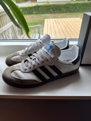 Adidas Samba vita och svarta sneakers - Klassiska Adidas Samba sneakers i vitt och svart med de ikoniska tre ränderna på sidan. Ovandelen är i läder med grå mockadetaljer vid tån och svart yttersula. Låg modell med snörning och blå Adidas-logga på plösen.