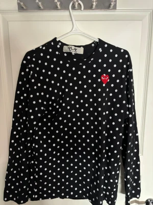 Commes des Garçons  - Svart långärmad tröja från Comme des Garçons med vita prickar och ett rött hjärta broderat på bröstet. Tillverkad i mjuk bomull och har en klassisk rund halsringning. Perfekt för dig som gillar lekfulla detaljer och stilren design. Är en XL men känns mer som en L/M till storlek.