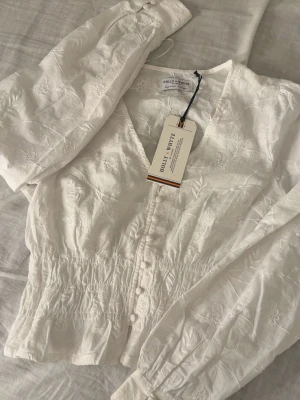 Vit blus från Holly & Whyte - Säljer en vit blus från Holly & Whyte med v-ringning, knappar framtill och smockad midja. Blusen har ett diskret blommönster i tyget och långa ärmar. Perfekt för en fräsch och stilren look.