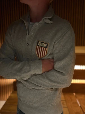 Grå collegetröja med emblem - Säljer en grå collegetröja med krage och knappar vid halsen. På bröstet sitter ett stort broderat emblem med texten '19 RLPC-34 United States' och röda ränder. Tröjan har fickor framtill och ribbade muddar vid ärmslut och nederkant. Perfekt till jeans för en avslappnad look.