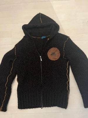 Mörkbrun stickad hoodie med dragkedja - Säljer en mörkbrun stickad hoodie med dragkedja och huva. Tröjan har ett stort brunt tygmärke på bröstet och dekorativa sömmar i kontrastfärg längs ärmar och sidor. Ribbstickade muddar vid ärmslut och nederkant ger en schysst passform.Hör av dig via frågor och funderingar 🙌😊