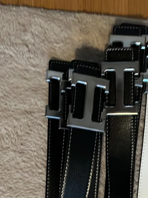 Ett svart bälte med silverfärgat spänne - Säljer ett svart bälte med bred design och stora, fyrkantiga silverfärgade spännen. Bältet har vita kontrastsömmar längs kanterna och är tillverkade i ett material som liknar skinn. Perfekt accessoar för att lyfta din outfit.