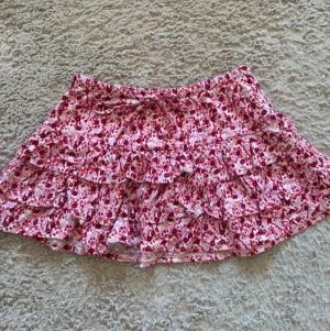Blommig volangkjol i rosa och vit - Söt kort kjol med blommigt mönster i rosa och vitt. Kjolen har flera volanger och en bekväm resår i midjan. Perfekt för varma dagar och ger en romantisk vibe till din outfit. Vill bli av med fort så pris kan diskuteras vid mycket snabb affär! 