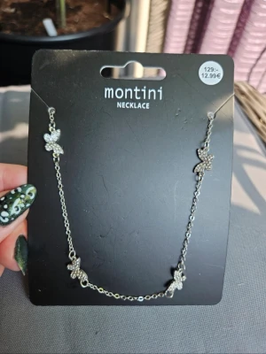 Silverfärgat halsband med fjärilar Montini - Snyggt silverfärgat halsband från Montini med fyra glittrande fjärilsberlocker. Kedjan är tunn och elegant, och fjärilarna är dekorerade med små gnistrande stenar som ger ett lyxigt intryck. Perfekt för att lyfta din outfit med en trendig detalj  Kan även hämtas I Partille  E3