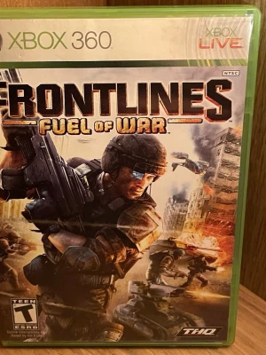 Frontlines: Fuel of War Xbox 360 spel - Frontlines: Fuel of War till Xbox 360 är ett actionfyllt förstapersonsskjutspel där du slåss i framtidens krigszoner. Omslaget har en soldat i stridsmiljö med explosioner och fordon. Spelet har stöd för multiplayer och stora kartor. Fodralet är grönt och spelet ges ut av THQ. Perfekt Skick