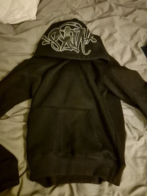 Svart hoodie från SNAK - Svart hoodie från SNAK med stor ficka framtill och grafittiliknande logga på huvan. Hoodien har en klassisk passform och är tillverkad i mjukt material som passar perfekt för chill dagar. Snygg streetwear vibe!