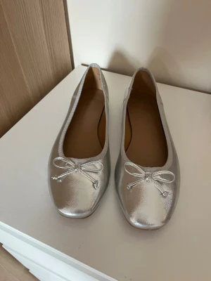Silvriga ballerinaskor med rosett - Snygga ballerinaskor i metallic silver från ASOS. Skorna har rund tå, dekorativ rosett framtill och en klassisk låg klack. Utsidan är i glansigt material och insidan är beige. Perfekta för dig som vill sticka ut lite extra.