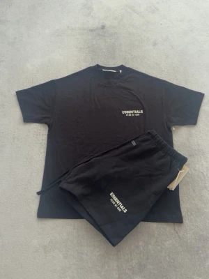 Svart set från Essentials Fear of God - Snyggt svart set med t-shirt och shorts från Essentials Fear of God. Båda plaggen har vit logga tryckt fram och bak. T-shirten har rund hals och korta ärmar, shortsen har resår i midjan och dragsko. Perfekt för en avslappnad och trendig look.
