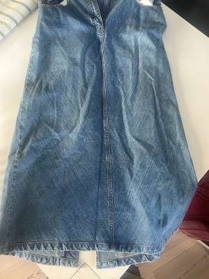 Blå jeanskjol med slits - Säljer en klassisk blå jeanskjol i rak modell med snygg slits baktill. Kjolen har en enkel design utan synliga detaljer och är gjord i ett tåligt denimtyg. Perfekt för dig som gillar en tidlös och avslappnad stil.