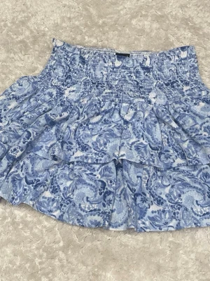 Blå volangkjol från Gina Tricot - Supersöt kort kjol från Gina Tricot i blått och vitt med volanger och smock i midjan. Kjolen har ett blommigt mönster och är gjord i mjuk bomull, perfekt för en fräsch och somrig look.