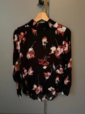 Svart blommig blus med hög krage - Snygg svart blus med röda och vita blommor och hög krage. Blusen har långa ärmar och är gjord i ett tunt, lätt material som ger ett luftigt intryck. Perfekt för dig som gillar en romantisk och trendig stil.