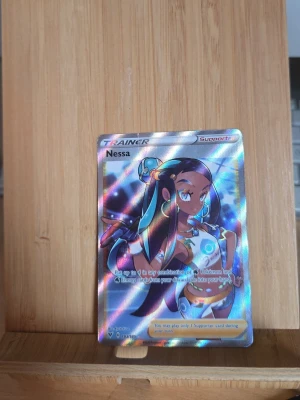 Pokémonkort Nessa Trainer holo - Säljer ett Pokémonkort av typen Trainer med motivet Nessa. Kortet är holografiskt med färgglada reflektioner och har texten 'Supporter' i hörnet. Perfekt för samlare eller Pokémon-fans som vill utöka sin kortsamling.