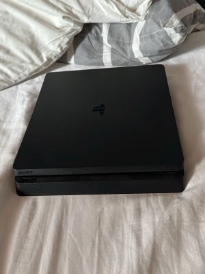 PlayStation 4 Slim spelkonsol svart - Säljer en svart PlayStation 4 Slim från Sony. Konsolen har en stilren, tunn och fyrkantig design med mattsvart finish och PlayStation-logga på ovansidan. Materialet är plast och metall. Perfekt för gaming och streaming.