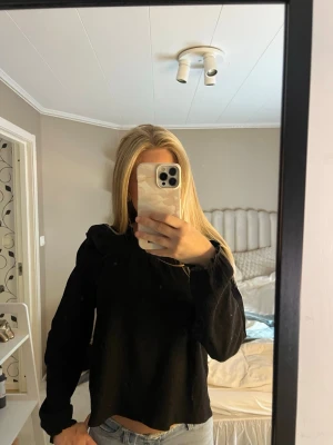 Svart blus med volang - Snygg svart blus med långa ärmar och volangdetaljer vid axlarna. Blusen har en rak passform och är tillverkad i ett mjukt, lätt material som ger en stilren look. Perfekt att matcha med jeans eller kjol för en trendig outfit.