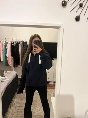 Russemerch från ginatricot - Mörkblå hoodie med dragkedja och vita snören i huvan. Klassisk modell med fickor framtill och ribbade muddar vid ärmslut och nederkant. Perfekt för en avslappnad och stilren look.