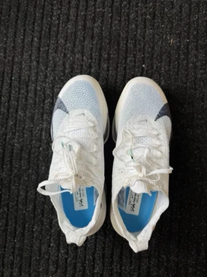 Vita Nike ZoomX sneakers - Säljer ett par vita Nike ZoomX sneakers med stickad ovandel och blå detaljer. Skorna har en låg profil, snörning och en tjock sula med synlig dämpning. Perfekta för dig som gillar sportig och modern stil.