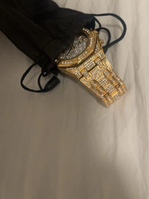 Prince klocka Iced-Out - Säljer en riktigt blingad klocka i guld med massor av glittrande stenar över hela boetten och armbandet. Klockan har en unik länkdesign och en rund urtavla som också är täckt av stenar. Perfekt för dig som vill sticka ut och gillar lyxig stil.