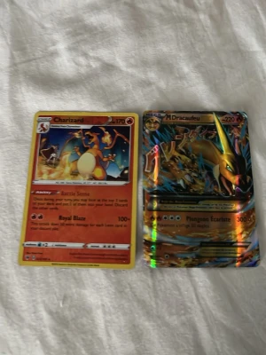 Pokémonkort Charizard & Dracaufeu - Två Pokémonkort: ett Charizard-kort med röd ram och ett glansigt M Dracaufeu EX-kort med färgstark design. Båda korten har motiv av eldpokémonen Charizard och är tryckta på kartong. Perfekt för samlare eller Pokémonfans.