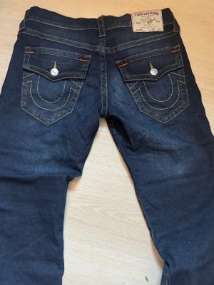  True Religion jeans - Säljer ett par mörkblå jeans från True Religion med kontrastsömmar och stora bakfickor med lock och dekorativa sömmar. Klassisk femficksmodell i denim med detaljer i orange och vita knappar. Perfekta för en trendig streetwear-look. Pris kan diskuteras💕