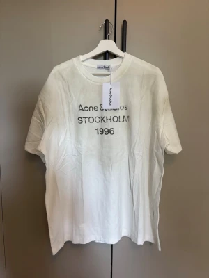 Vit Acne Studios t-shirt Stockholm - Vit t-shirt från Acne Studios med trycket 'Acne Studios STOCKHOLM 1996' på bröstet. Klassisk passform med korta ärmar och rund hals. Tillverkad i 100% bomull för en skön och luftig känsla. Perfekt för en clean och stilren look.