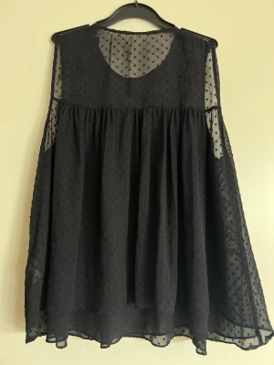 Svart broderad klänning med prickar - Säljer en svart blus med transparenta partier, broderade blommor på bröstet och små prickar i tyget. Blusen har en lös passform och är ärmlös, perfekt för dig som gillar detaljer och en luftig känsla. Lappen är borttagen i nacken. Storleken är S men passar också M. Längd: 65 cm ca. 