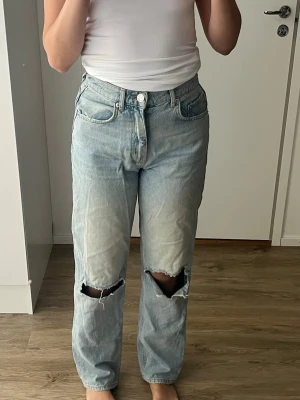 Ljusblå vida jeans med hål på knäna - Säljer ett par ljusblå vida jeans med stora slitningar över båda knäna. Jeansen har hög midja, klassisk femficksmodell och stängs med knapp och dragkedja. 