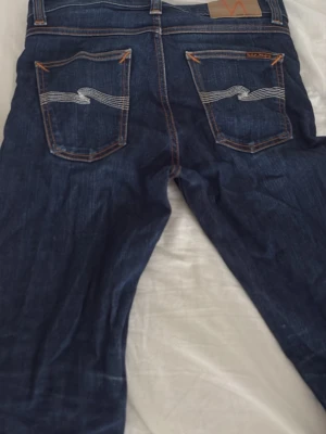 Mörkblå raka jeans från Nudie Jeans - Snygga mörkblå jeans från Nudie Jeans med klassisk femficksmodell och kontrastsömmar. Jeansen har rak passform och är tillverkade i slitstarkt denimtyg. Perfekta för dig som gillar en tidlös och clean look.