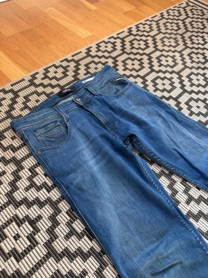 Replay Anbass - Säljer ett par blå slim fit jeans från Replay i modellen ”Anbass” med klassisk femficksdesign och dragkedja. Jeansen har snygga slitningar och är tillverkade i stretchigt denimtyg för skön passform. Storleken är W32L32