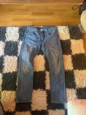 Blå raka jeans i klassisk modell - Säljer ett par blå jeans med rak passform och fem fickor. Jeansen har normal midja och klassisk denimlook med ljus tvätt. Materialet är bomullsdenim och de har bälteshällor samt knappgylf. Perfekta för en avslappnad stil.
