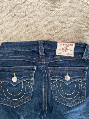True Religion blå jeans med fickdetaljer - Säljer ett par blå jeans från True Religion med ikoniska kontrastsömmar och stora fickor bak med lock och silverknappar. Klassisk patch med logga bak i midjan. Snygg passform och coola detaljer som sticker ut.