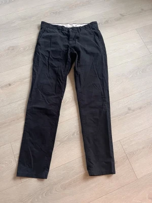 Svarta slim fit chinos från H&M - Snygga svarta chinos från H&M:s L.O.G.G.-kollektion i slim fit-modell. Byxorna har klassisk design med fickor fram och bak samt bälteshällor. Tillverkade i mjukt bomullstyg som ger en skön känsla och stilren look. Den ena knappen på bakfickan saknas, annars i fint skick. Storlek storlek 34 herr.