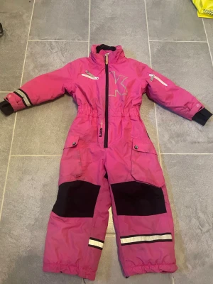 Rosa overall med svarta detaljer - En rosa overall med svarta förstärkningar på knän och bak, reflexdetaljer på ben och ärmar samt dragkedja framtill. Overallen har elastisk midja, fickor med knappar och muddar vid ärmslut. Perfekt för kalla dagar och utomhuslek.
