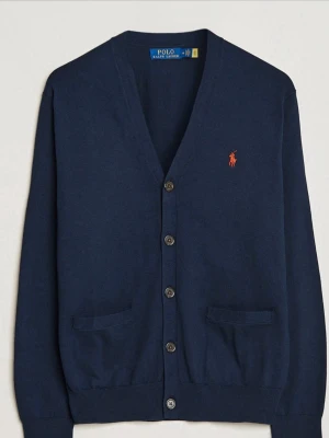 Marinblå cardigan från Polo Ralph Lauren - Klassisk marinblå cardigan från Polo Ralph Lauren med v-ringning, knappar framtill och två fickor. Tröjan har lång ärm och den ikoniska röda loggan broderad på bröstet. Perfekt att slänga över en t-shirt eller skjorta.