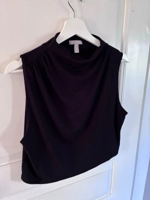 Svart ärmlös topp från H&M - Snygg svart ärmlös topp från H&M med hög hals och avslappnad passform. Perfekt att styla med jeans eller kjol för en clean och modern look. Tillverkad i mjukt material som känns skönt mot huden.