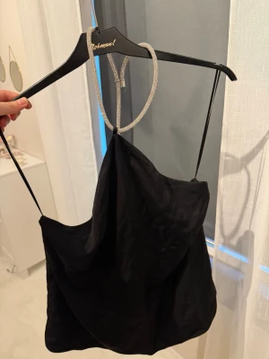 Svart halternecktopp med strassband - Snyggt svart halternecktopp  i satin med tunna axelband och ett glittrande strassband som går runt halsen. Toppen har en lös passform och draperad framsida som ger en trendig look. Perfekt för att lyfta vilken outfit som helst.