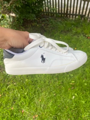 Vita Polo Ralph Lauren Theron V sneakers stl 35 - Fräscha vita sneakers från Polo Ralph Lauren med klassisk svart logga på sidan och svart detalj med 'POLO' på hälen. Skorna har vita snören och är tillverkade i slätt syntetmaterial med låg profil. Perfekta för en clean och stilren look.