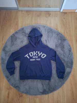 Mörkblå hoodie med Tokyo-tryck - Mörkblå hoodie med stor huva och magficka. På bröstet står det 'TOKYO JAPAN MOUNT FUJI' i vitt tryck med blockbokstäver. Hoodien har ribbade muddar vid ärmar och nederkant. Perfekt för en avslappnad streetwear-look.